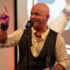 Keynote speaker Kevin Bulmer pointing a finger upward on stage with a microphone, emphasizing a critical point about the subconscious mind and positive language.