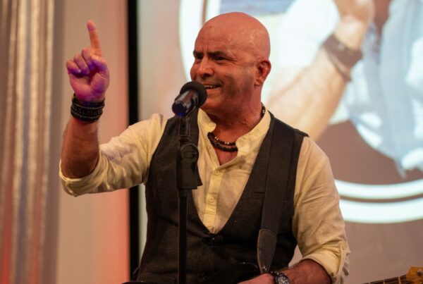 Keynote speaker Kevin Bulmer pointing a finger upward on stage with a microphone, emphasizing a critical point about the subconscious mind and positive language.