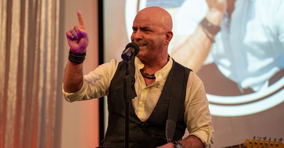 Keynote speaker Kevin Bulmer pointing a finger upward on stage with a microphone, emphasizing a critical point about the subconscious mind and positive language.