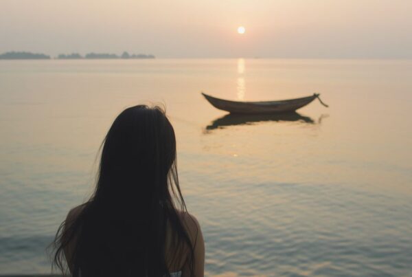 A young woman sitting on a shoreline looking at an expansive body of water with a small wooden boat drifting nearby at sunset, representing self-reflection and recalibrating one's path.