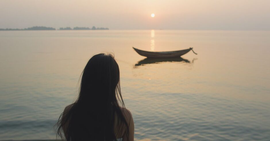 A young woman sitting on a shoreline looking at an expansive body of water with a small wooden boat drifting nearby at sunset, representing self-reflection and recalibrating one's path.