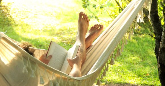 A person lounging in a hammock on a sunny summer day, feet crossed and relaxed, reading a book with lush green trees in the background.