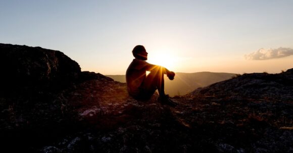 A person sitting alone on a mountain peak at sunset, gazing out over the landscape in quiet reflection or meditation, with warm golden light filling the sky.