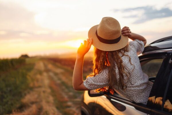 Young woman with her head out the car window, riding into a golden sunset — a visual blend of restful escape and adventurous freedom.
