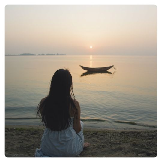 A woman viewed from behind as she gazes out at a calm lake or ocean at sundown where a small boat sits adrift on the water, representing the season of feeling off course and the invitation to realign with what matters most through the REAL Success Crew membership.